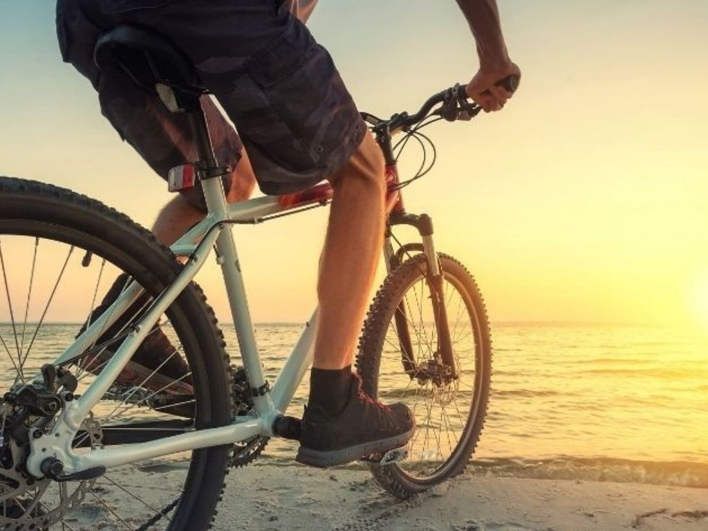 A close up photo showing the bottom half of a man's shorts and legs on a bike. He is riding along the coastline as the sun starts to go down.