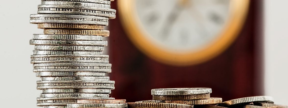 A stack of silver coins with a burgundy wall and a round wall clock trimmed in gold blurred out in the background. This photo indicates the importance of budgeting for Furniture decorating.