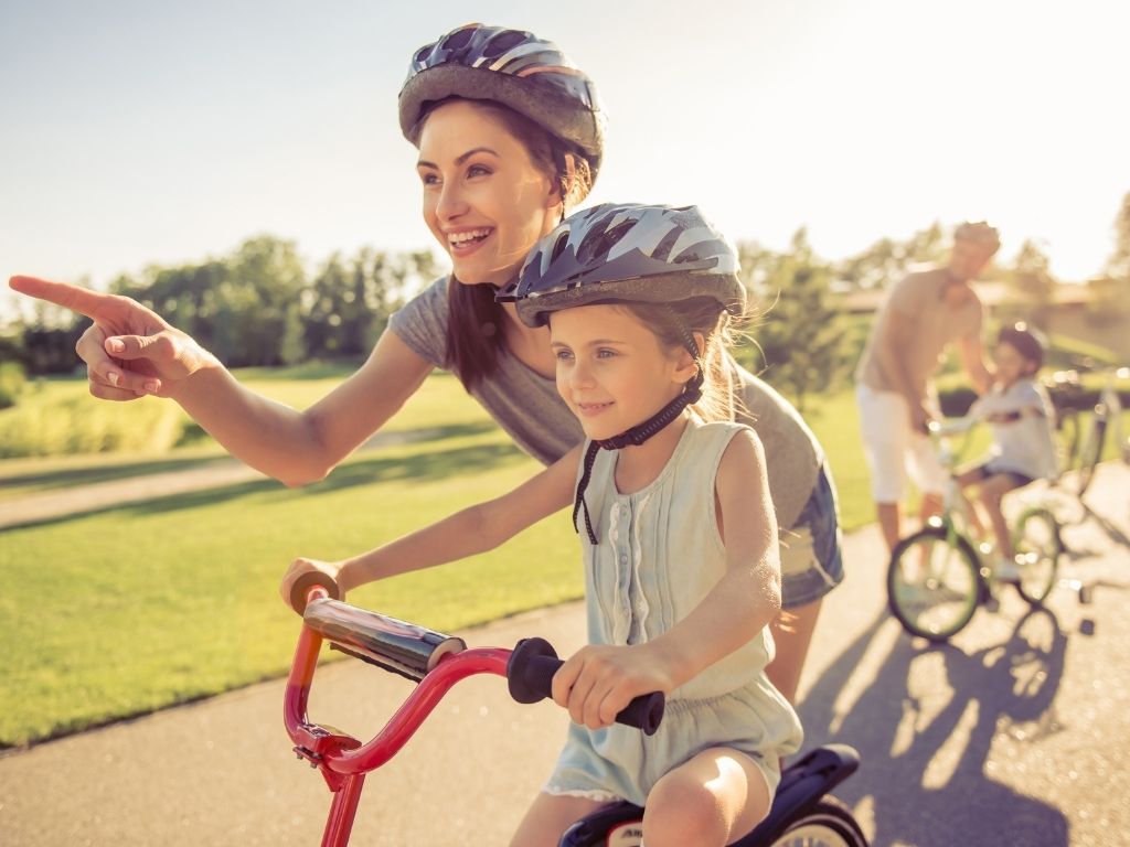 A mom in a bike helmet. A young daughter around the age of 7 on a red bike wearing a helmet. The mom is helping the daughter learn to ride.