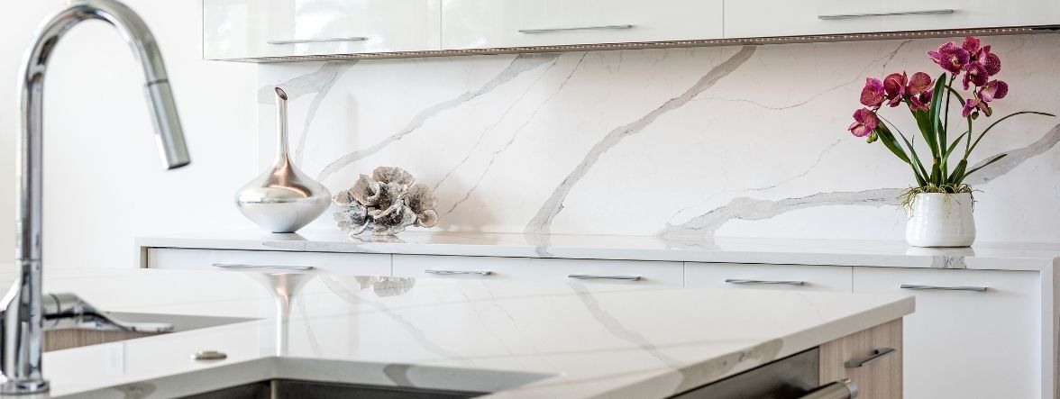 A close up of a contemporary style kitchen showing the use of colors for furniture decorating. The main color is white and is used in the upper cabinets and the walls. The backsplash is a white background with gray veining, pull in the second color for the room.  The third color is a pop of pink in the flowers set upon the kitchen coutertop. 