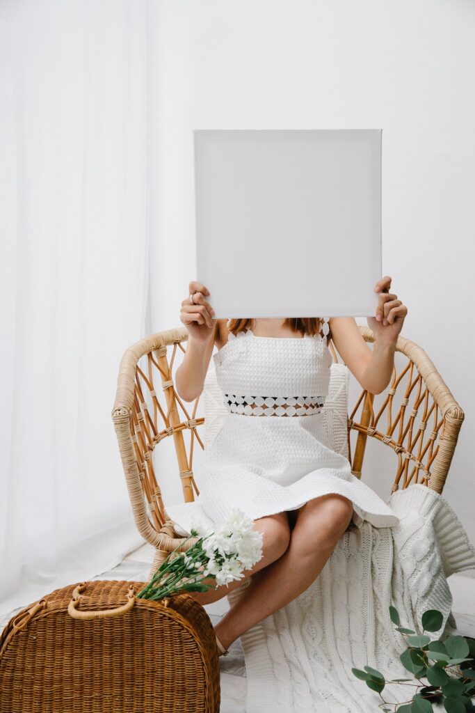A photo of a girl sitting in a wicker chair wearing a white dress and holding a large white painted square in front of her face. The photo shows how white is one of the paint colors to help cool a home.