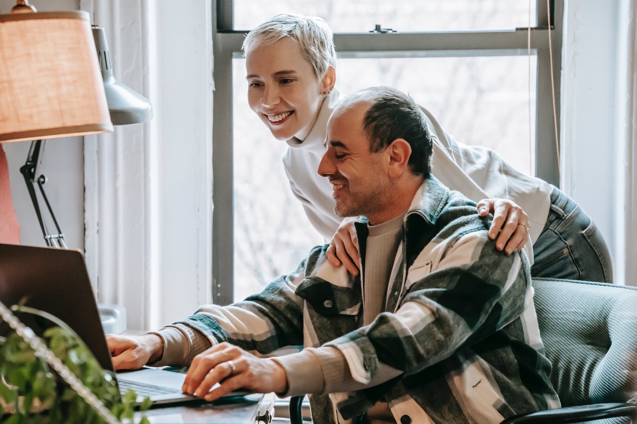 A middle-aged woman with silver hair is leaning forward looking at a laptop with her hands on her husband's shoulders. Her husband is wearing a plaid green shirt and sitting in a chair. He is also looking at the laptop, browsing for 55+ communities in Longboat Key.