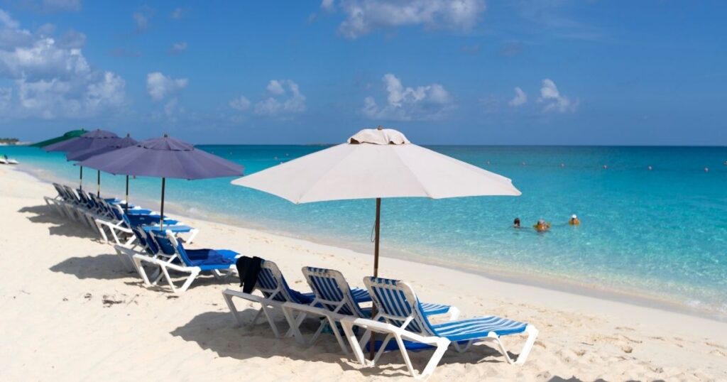 A picture of the beach showing the soft white sand and crystal blue water at Siesta Key Beach. Along the shore are blue and white striped beach chairs with some white and some blue umbrellas. Siesta Key beach is one of the most relaxing things to do in Sarasota.