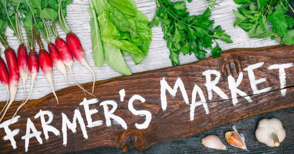 A rustic piece of plank wood with white painted letters spelling out Farmer's Market is laid on a table in front of a row of vegetables including radish, lettuce, parsley and mint.