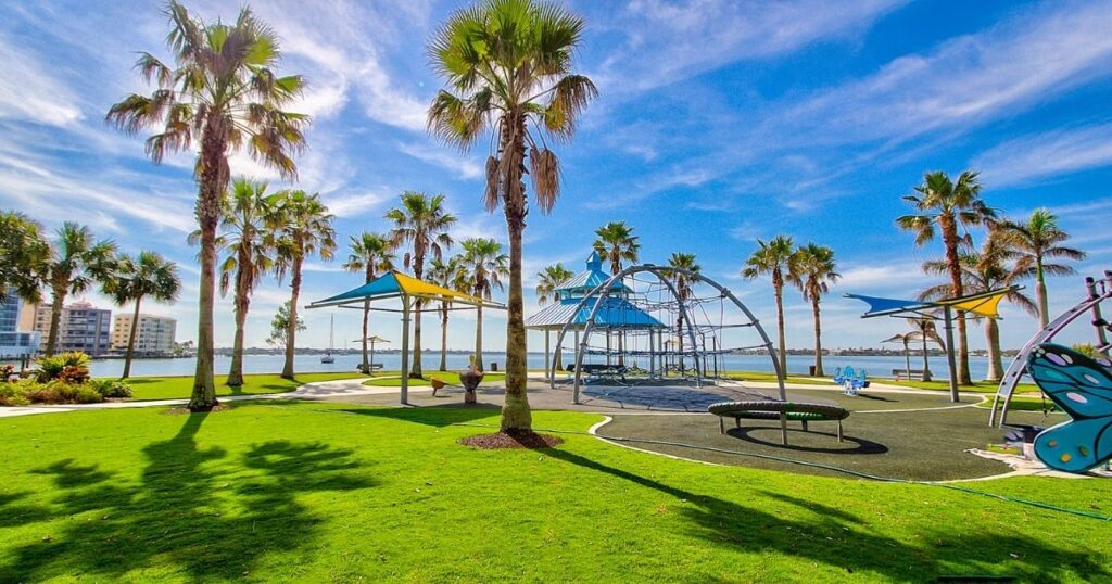 A photo of Harts Landing in Bayfront Park shows a green grassy area with an irregular shape sand playground area. There is a blue roof pavilion, and some of the playground equipment has bright blue and yellow sail cloths over them to protect children from the sun.