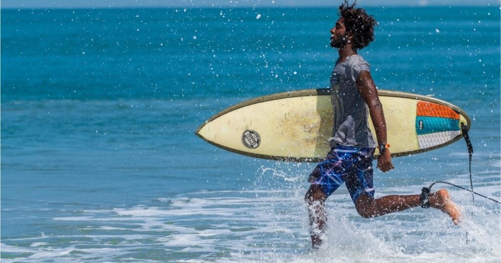 A photo showing surfing as one of the things to do in Sarasota and Longboat Key. The young man is in blue shorts carrying a surfboard running along the edge of the water.